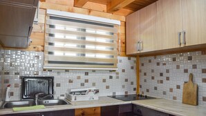 Une cuisine avec des meubles de rangement en bois, un pare-feu en carrelage, une fenêtre avec des stores et une planche à découper posée sur le plan de travail.