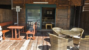 Une terrasse extérieure en bois, avec une table et des chaises en osier, un banc et une porte donnant sur un espace intérieur.
