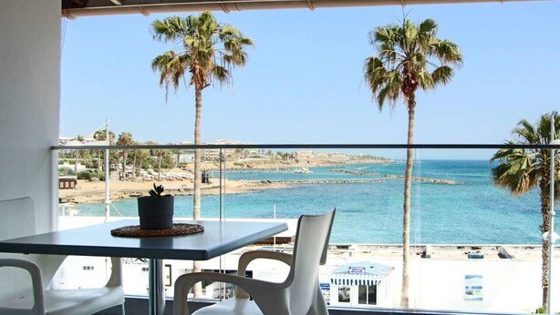 A balcony with a table and chairs overlooking a beach with palm trees and the sea. - Phaedrus Living: Seaview Luxury Flat Lighthouse 53 (Paphos)