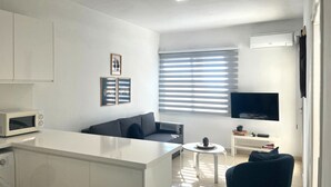Una sala de estar moderna con un sofá, una mesa de centro y un televisor. Cuenta con una zona de cocina con microondas y fregadero.