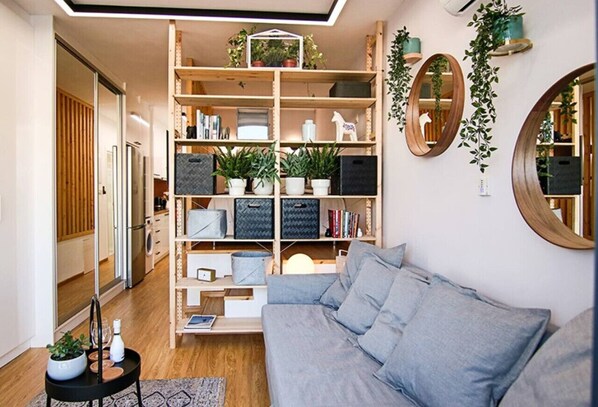 Una sala de estar moderna con un sofá gris, estanterías de madera con plantas y un espejo grande.