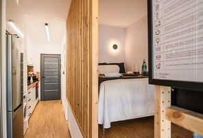 Una habitación de hotel moderna con un separador de madera, una cama con ropa blanca, una mesita con una botella y vasos, y un panel informativo en la pared.