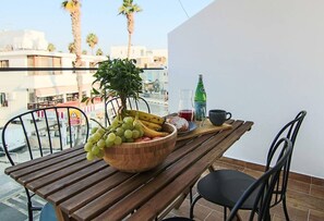 Una mesa de madera con un cuenco de fruta, una planta en maceta y una botella de agua en un balcón que ofrece vistas a palmeras y edificios.