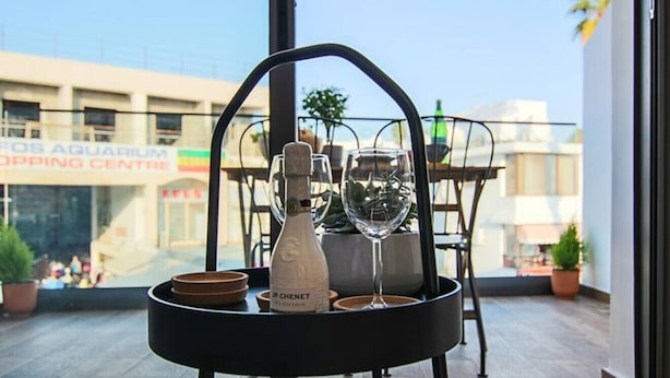 Una mesa de comedor con una botella de vino Chennet, dos copas de vino y una bandeja con cuencos, sobre un balcón con vistas a un centro comercial.
