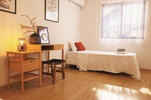 Une petite pièce bien éclairée, avec un bureau en bois, une chaise, un lit recouvert d’une courtepointe blanche et une fenêtre ornée de rideaux légers.