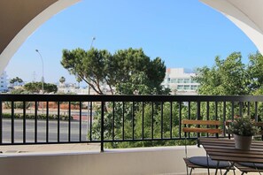 Un balcon avec un banc en bois et un pot de fleurs, donnant sur une rue bordée d’arbres et de bâtiments.