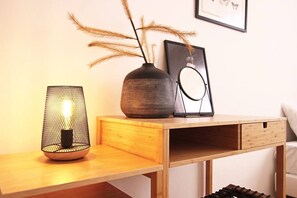 Ein Holztisch mit einer gewobenen Lampe, einer Vase mit Trockenpflanzen und einem gerahmten Bild an der Wand.