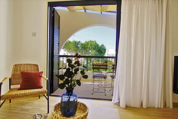 Ein Zimmer mit einem Korbstuhl, einem roten Kissen, einer Vase mit einer Pflanze und Blick auf einen Außenbalkon mit Geländer und einer Bank.