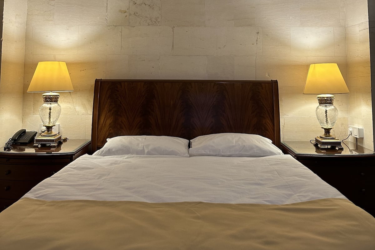 Een hotelkamer met een grote gewelfde stenen muur, een houten hoofdeinde, twee nachtkastjes met lampen en een net opgemaakt bed.
