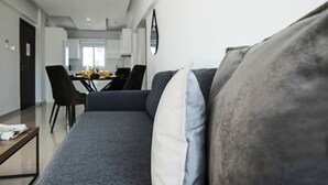 Moderni olohuone, jossa on harmaa sohva, taustalla ruokailutila ja avoimen oven kautta näkyvä keittiö.