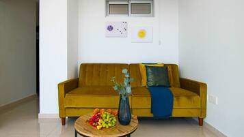 A living room with a yellow sofa, a round wooden coffee table with fruits and a vase, and two wall-mounted artworks.