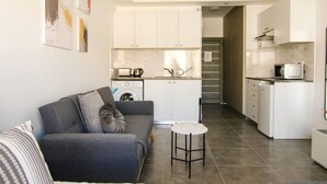 En moderne lejlighed med en grå sofa, et lille rundt bord, en vaskemaskine og et køkken med hvide skabe og en mikrobølgeovn.