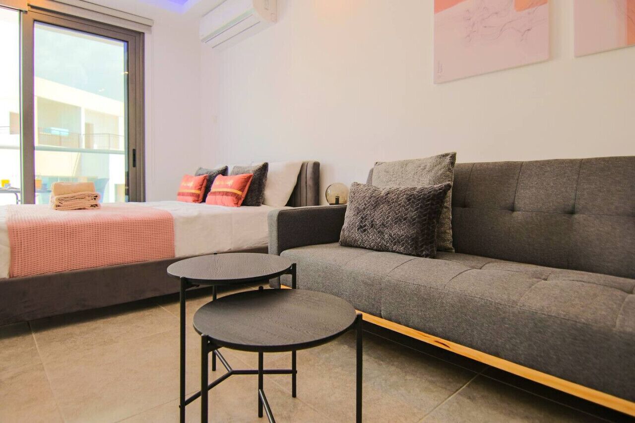 A modern living room with a grey sofa, two round tables, and a bed with a pink blanket. - Phaedrus Living: Seaside Luxury Flat Athina 115 (Paphos)