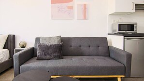 A modern living room with a grey sofa, a small round table, and a kitchen area with a microwave and refrigerator. - Phaedrus Living: Seaside Luxury Flat Athina 115 (Paphos)
