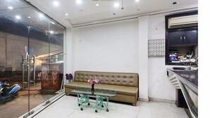 A modern living room with a glass partition, a brown sofa, a glass coffee table with flowers, and a kitchenette area. - Hotel Sunrise Dx @ Opposite New Delhi Railway Station (New Delhi)
