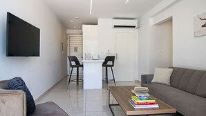 Un soggiorno moderno con un divano, un tavolino da caffè con libri, una TV a parete e un angolo cottura con un tavolo da pranzo e sedie.