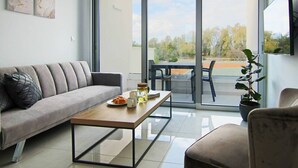 Ein modernes Wohnzimmer mit einer Couch, einem Couchtisch und einem Esstisch. Ein Glastür verschiebt sich zu einem Außenbereich mit Blick auf Bäume und einem Tennisplatz.