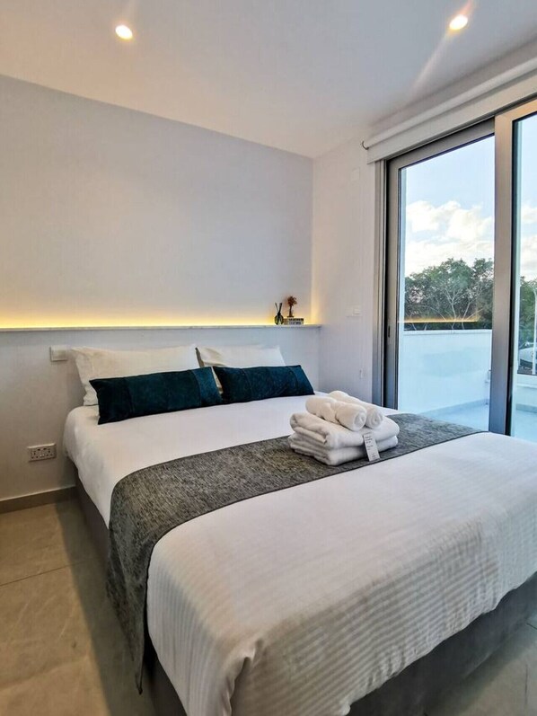 Habitación de hotel moderna con una cama grande, una mesita de noche con un florero y vistas al exterior a través de un ventanal amplio.