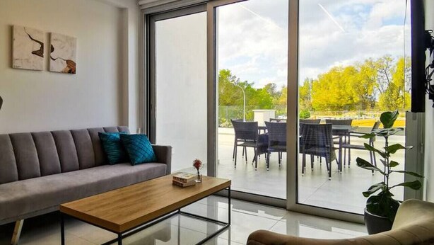 Een moderne woonkamer met een bank, salontafel en een fauteuil. Er is een groot schuifglasdeur die leidt naar een terras met een tafel en stoelen.