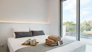 A modern hotel room with a large bed, a bedside table with a tray of cups and a towel, and a view of a landscaped area through a large window.