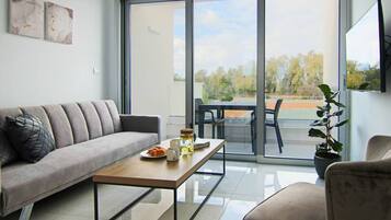 Sala de estar moderna com sofá, mesa de centro e mesa de jantar. Há uma porta de vidro deslizante que dá para área externa com vista para árvores e quadra de tênis.