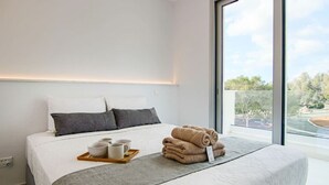 A modern hotel room with a large bed, a bedside table with a tray of breakfast items, and a view of a landscaped area through a large window. - Phaedrus Living Seaside Executive Flat Harbour 204 (Paphos)