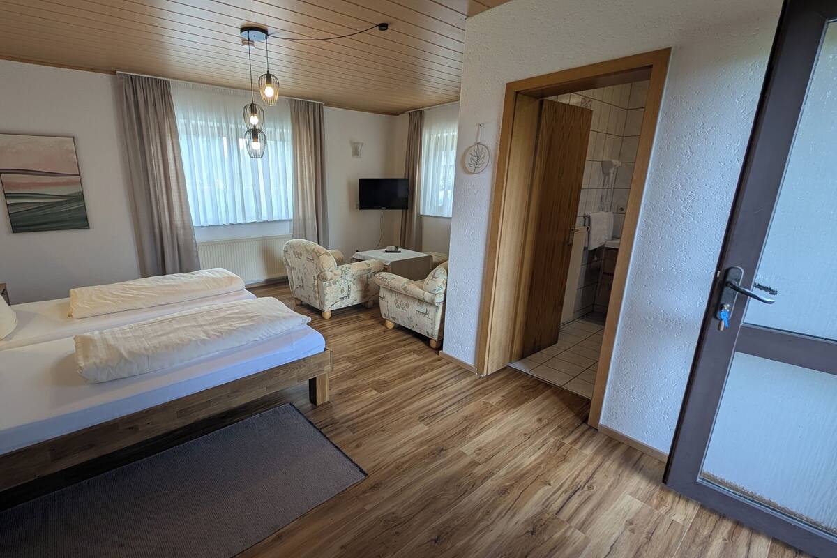 A room with a bed, two chairs, a TV, and a wooden floor.