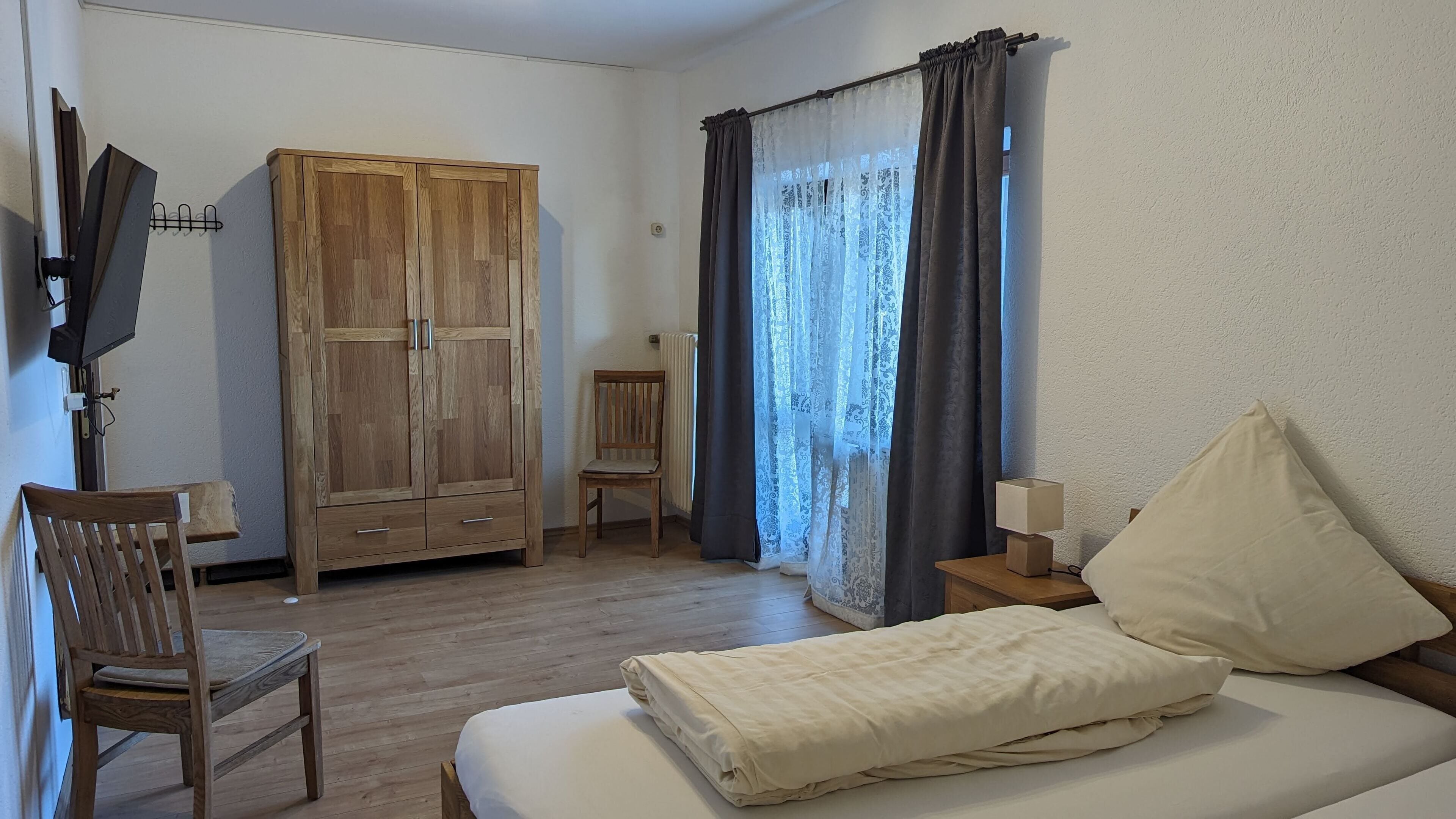 A bedroom with a wooden wardrobe, a chair, a bed with a white duvet, a nightstand with a lamp, and a window with curtains.