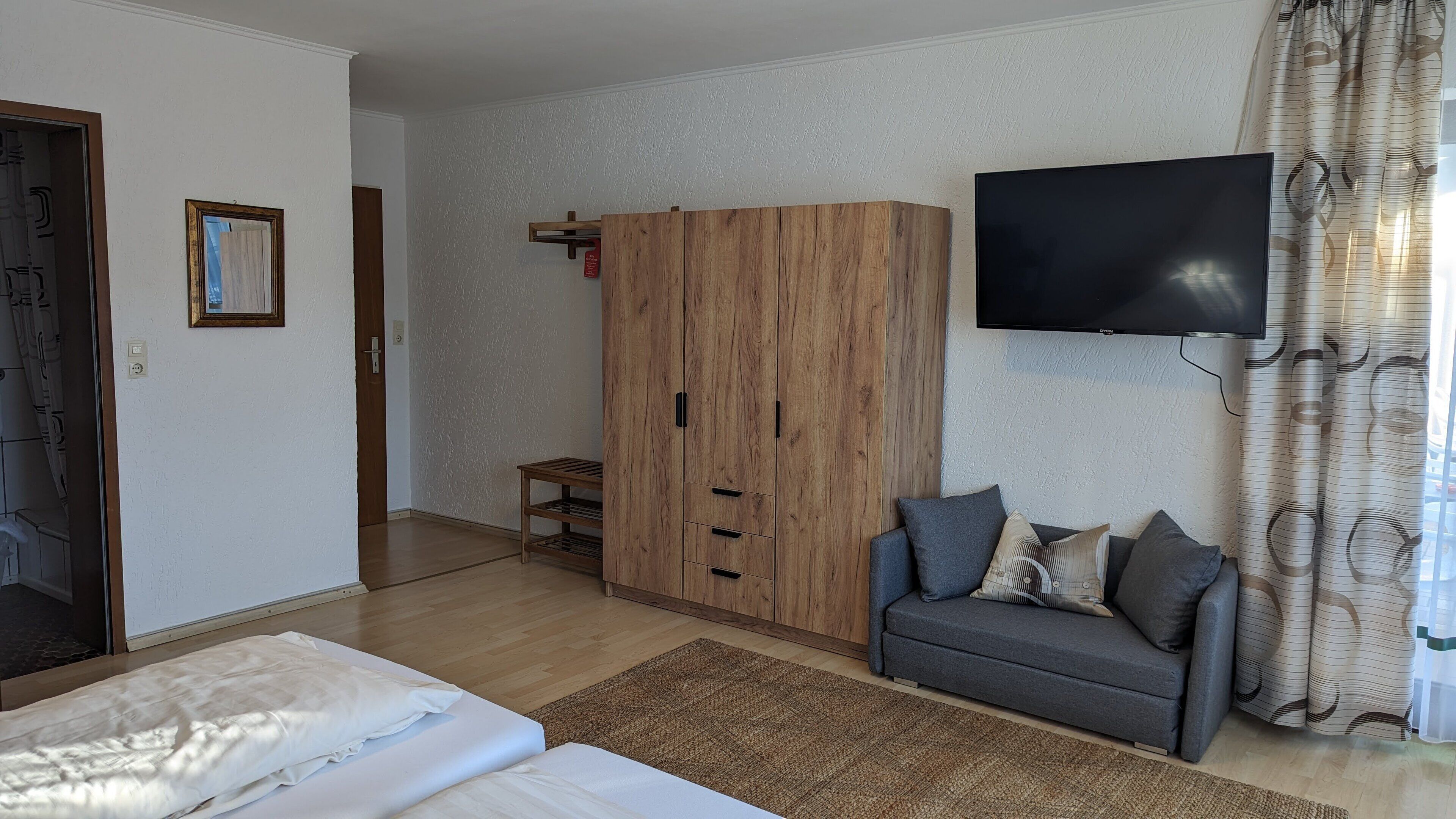 Ein Zimmer mit einem Bett, einem grauen Sofa, einem Holzschrank, einem Fernseher und einem gemusterten Vorhang.