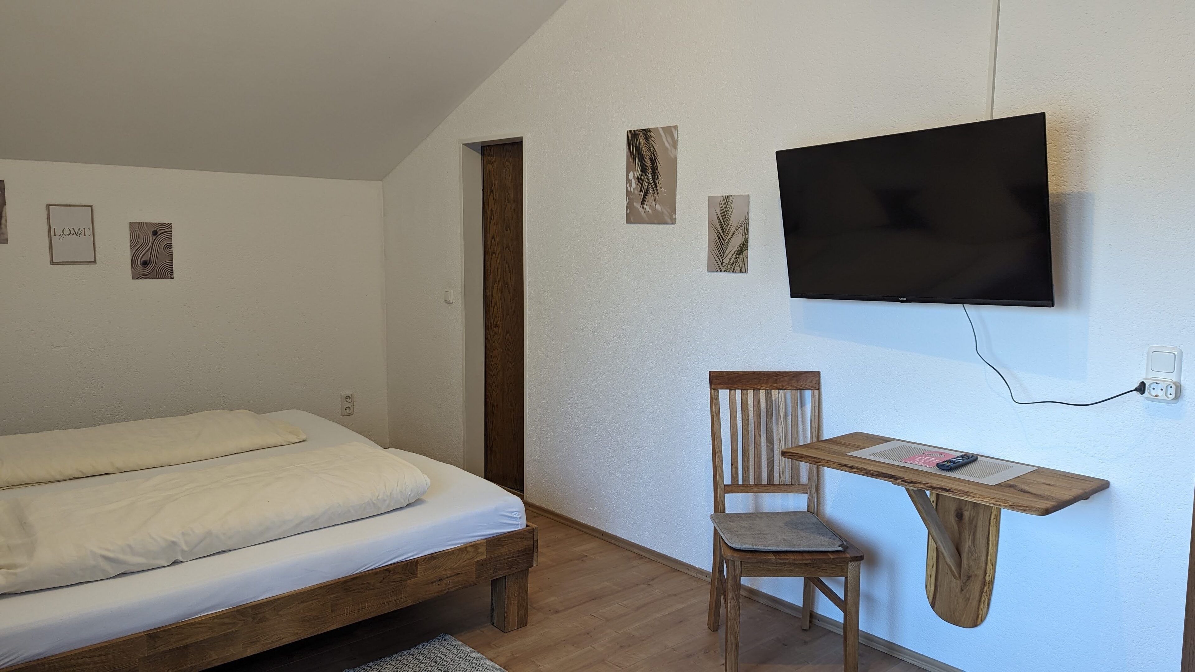 Un dormitorio con cama de madera, un escritorio con silla, un televisor de pantalla plana montado en la pared y una puerta.