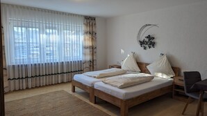 Un dormitorio con dos camas de madera, un ventanal con cortinas de gasa, un adorno en la pared y una mesita de noche de madera.
