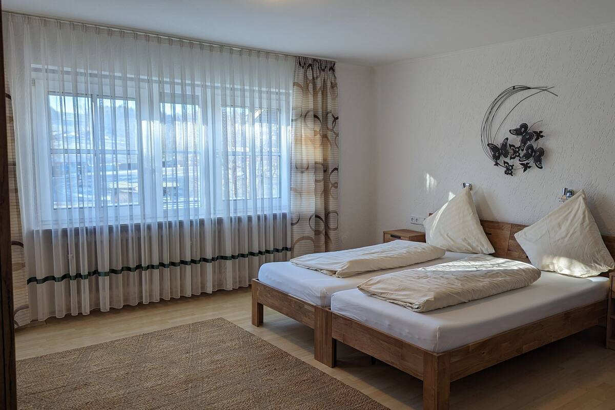 A bedroom with two wooden beds, a large window with sheer curtains, a decorative wall piece, and a wooden nightstand.