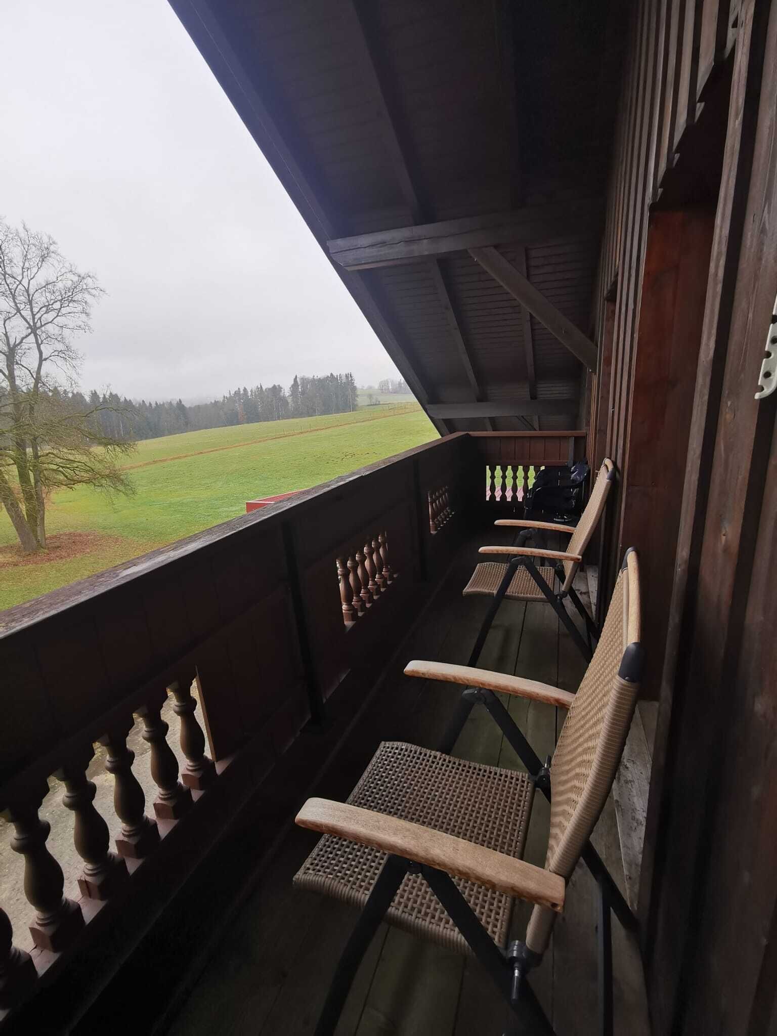 Un balcon en bois avec des chaises en osier, une vue sur un champ verdoyant et un ciel nuageux.