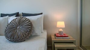 Un lit bien fait, agrémenté d’un coussin décoratif, d’une lampe de chevet et d’une pile de livres posée sur une petite table de chevet en bois.