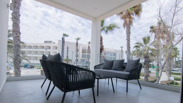 Een balkon met twee zwarte wicker stoelen en een grijze bank, met uitzicht op een parkeerplaats en gebouwen met palmbomen.