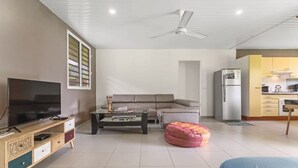 Un salon moderne doté d’un téléviseur à écran plat, d’un canapé, d’une table basse, d’un pouf, d’un ventilateur de plafond et d’un coin cuisine.