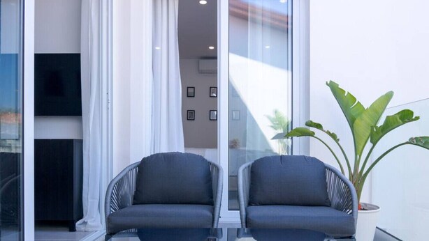 A modern room with two grey upholstered chairs, a large potted plant, and a glass table. - Phaedrus Living: Limnaria Luxury Flat Ivy 102 (Paphos)