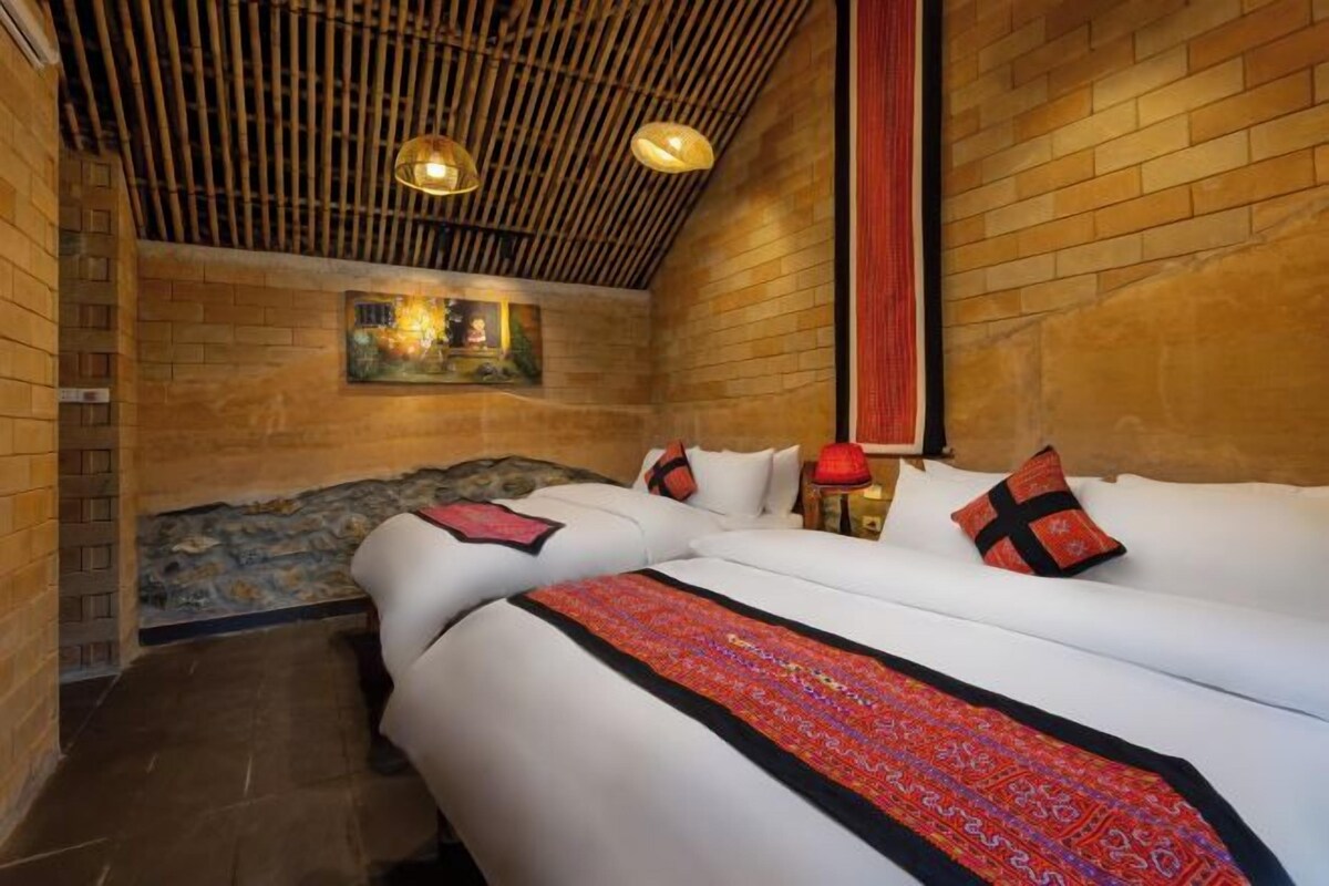 A room with a brick wall, a bed with white bedding and red accents, a wooden ceiling, and a small table with a lamp.