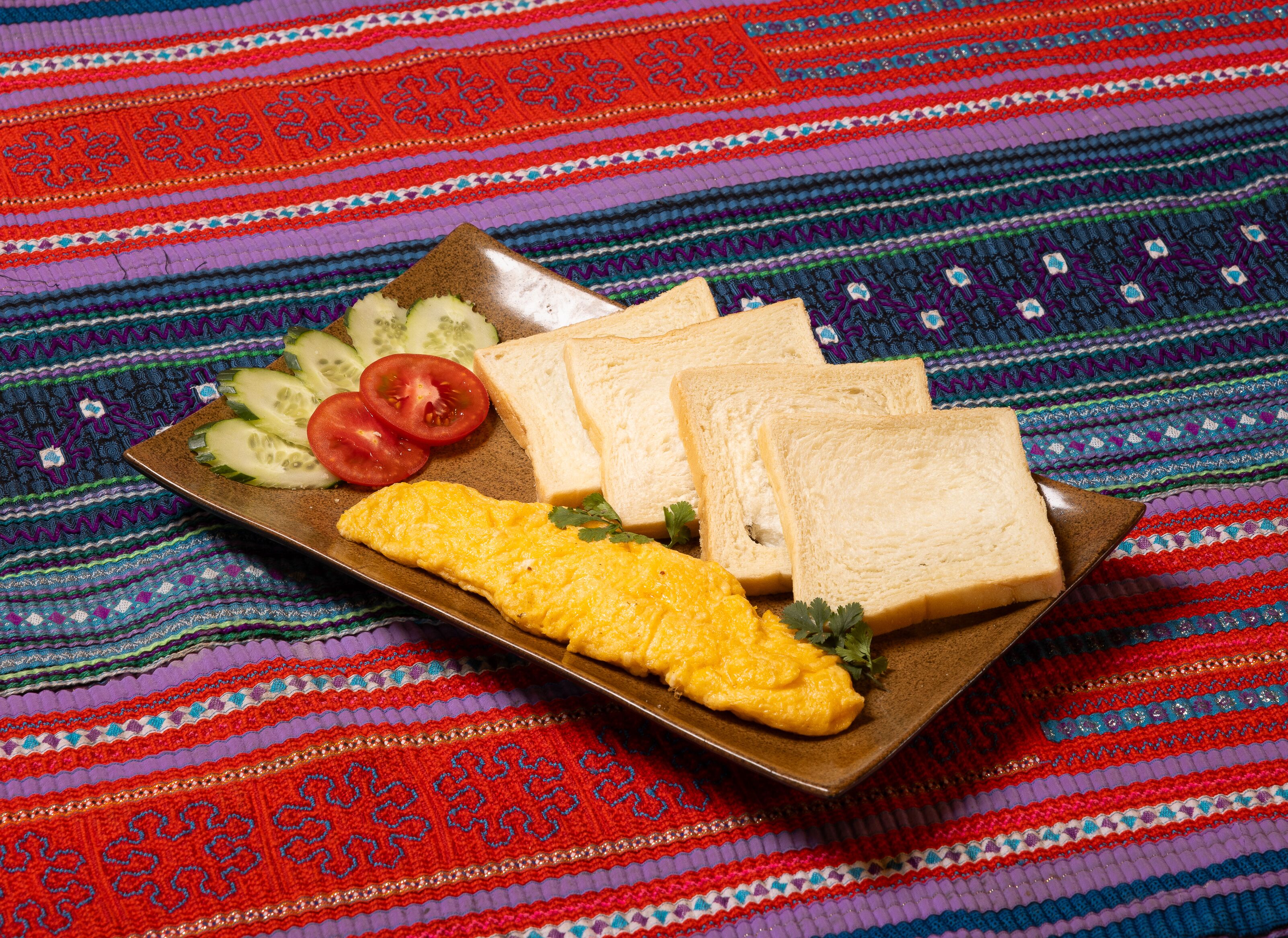 A plate with scrambled eggs, slices of tomato, cucumber, and two slices of white bread on a colorful woven tablecloth.