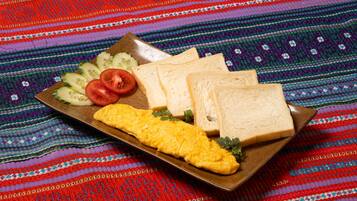 A plate with scrambled eggs, slices of tomato, cucumber, and two slices of white bread on a colorful woven tablecloth.