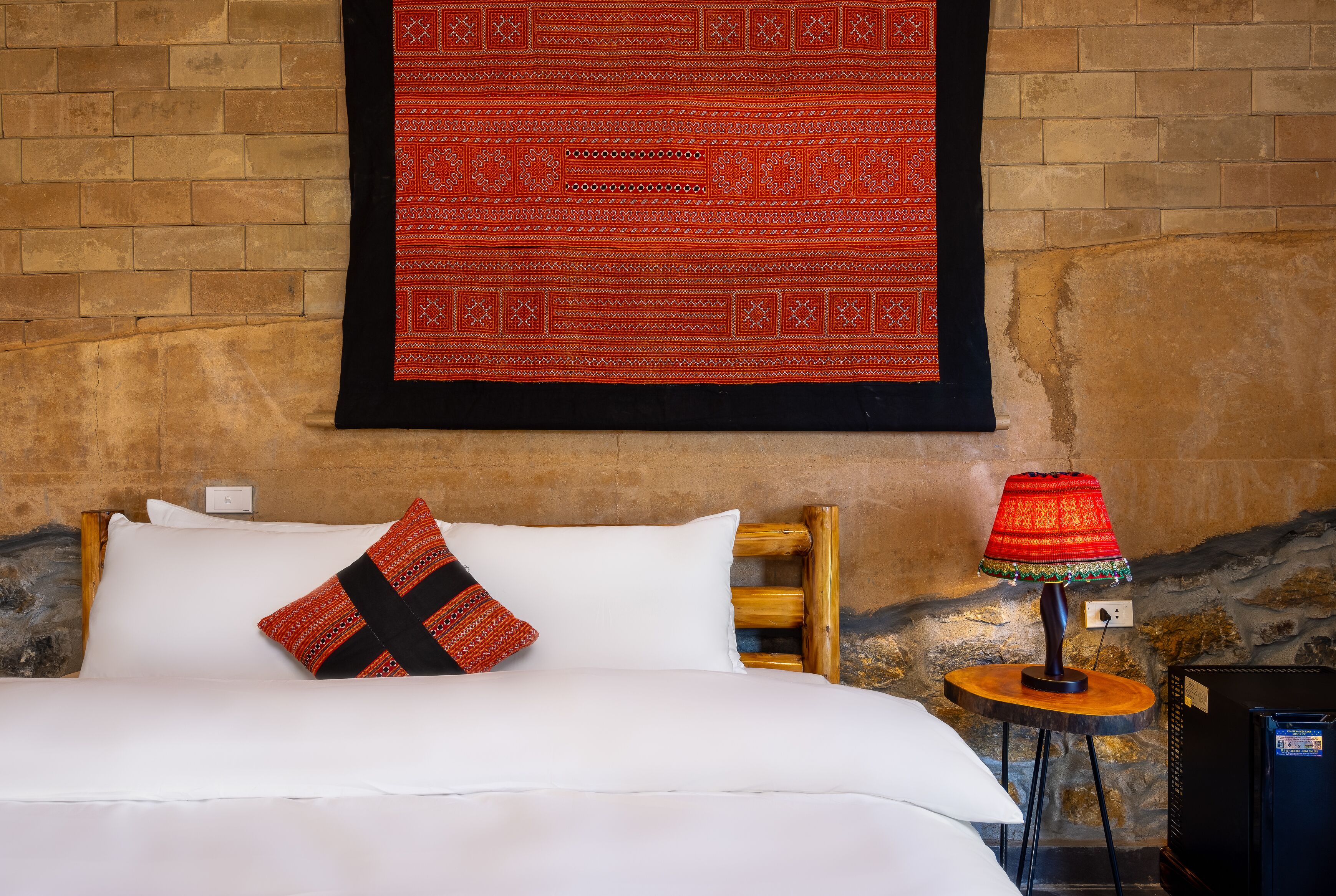 A bed with white bedding and a red and black patterned pillow, a red and gold lamp on a wooden table, and a red patterned tapestry on the wall.