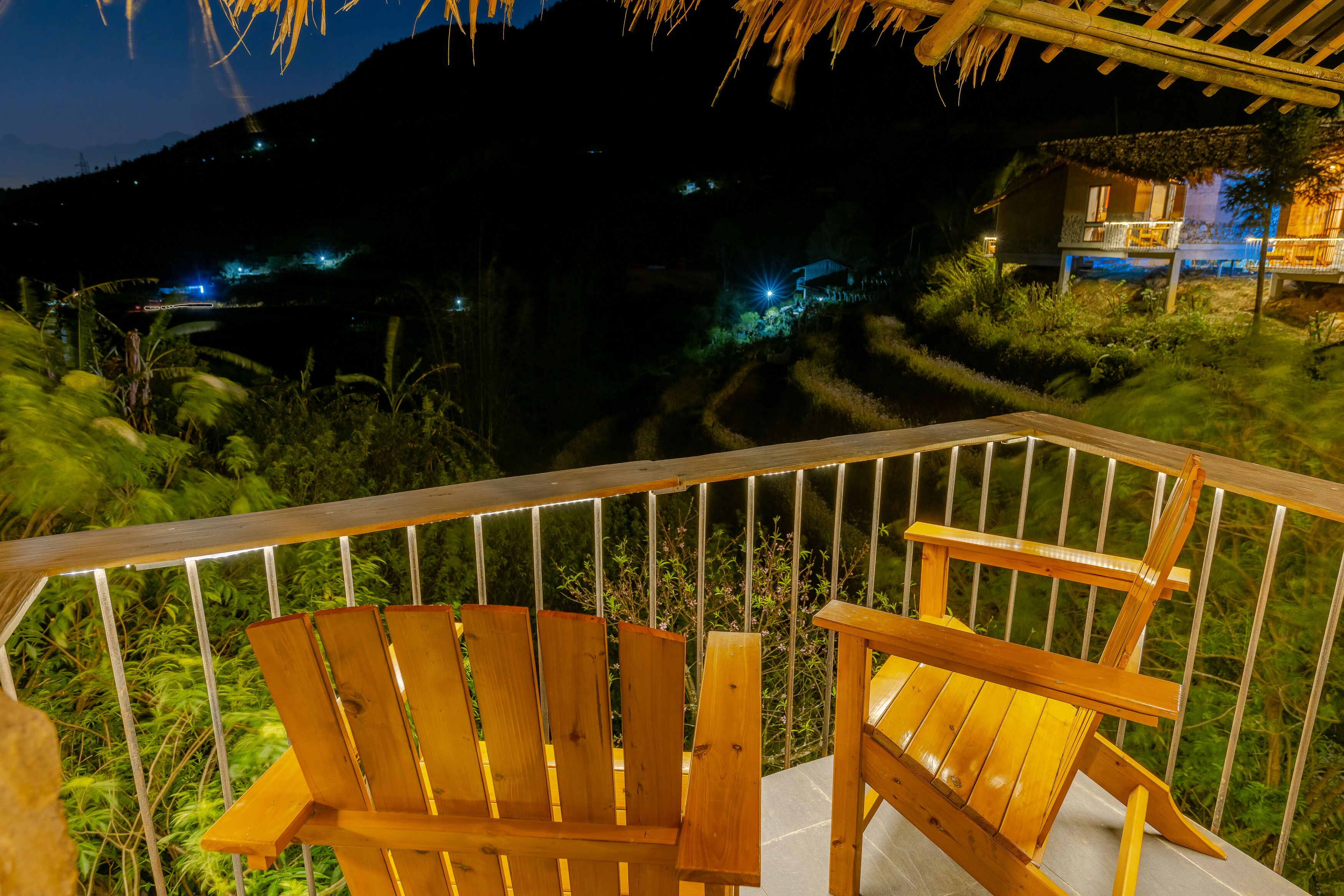 Una terraza de madera con dos sillas, con vistas a un paisaje escalonado y un edificio iluminado a lo lejos.