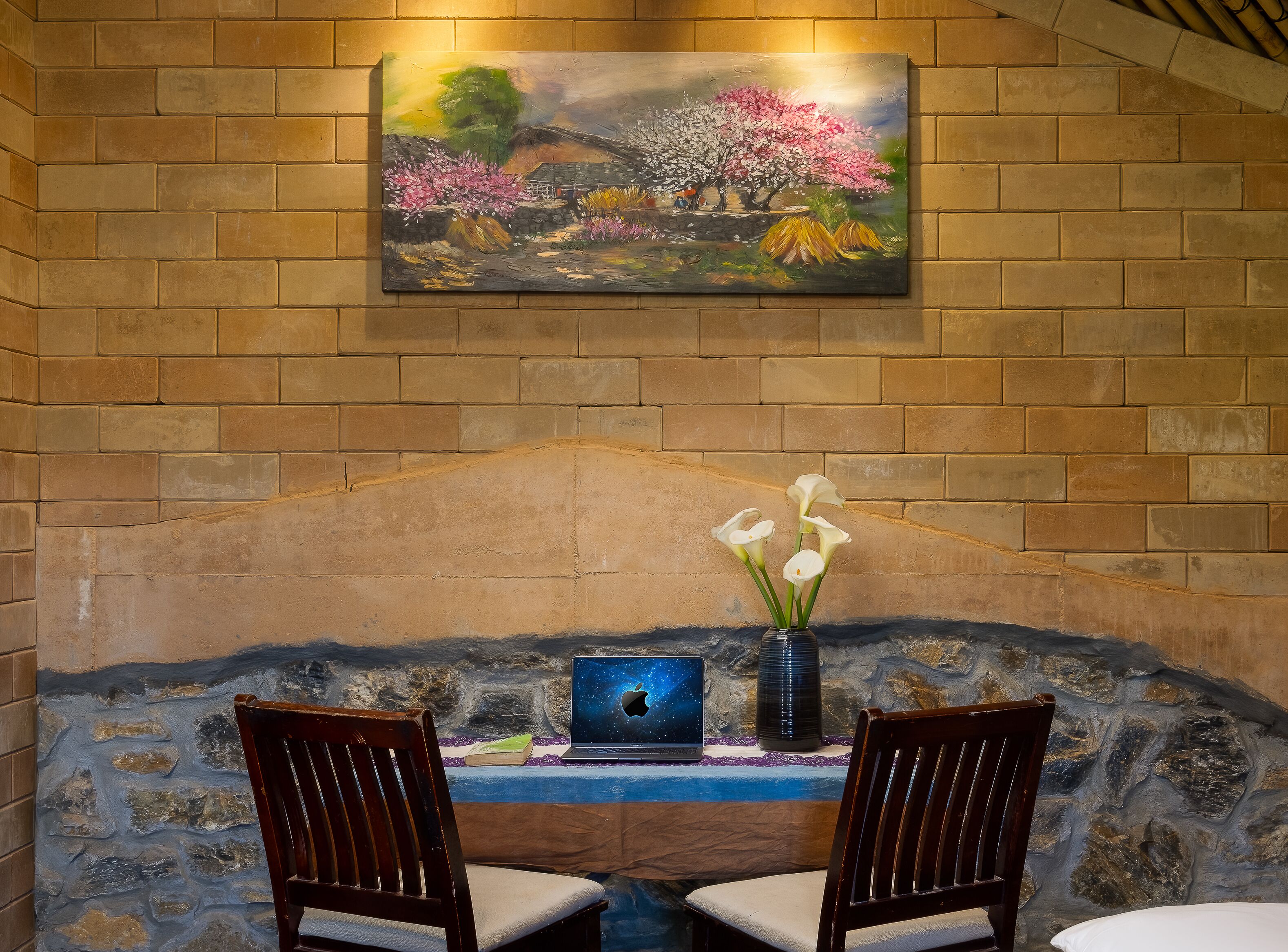 A room with a stone wall, a painting of a landscape, a laptop, and two chairs.