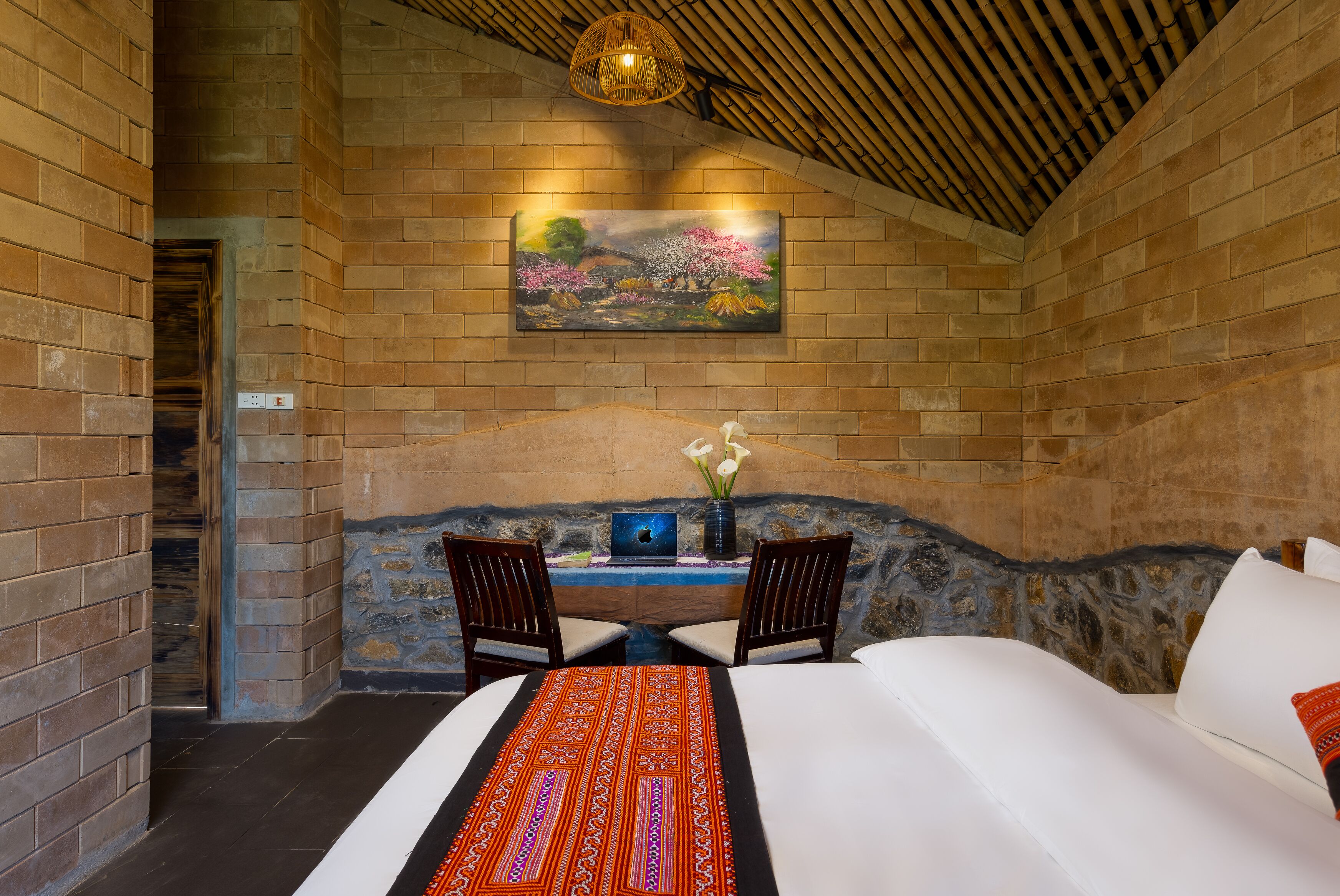 A room with a bed, two chairs, a table with a vase of flowers, and a painting on the wall.