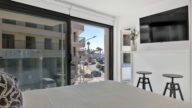 Una habitación de hotel moderna con una cama grande, un televisor montado en la pared y un balcón con vistas a la calle y edificios.