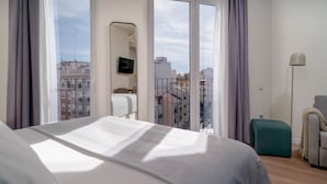Una habitación de hotel moderna con una cama grande, vistas de la ciudad a través de ventanas del suelo al techo, una pequeña zona de estar y un televisor montado en la pared.