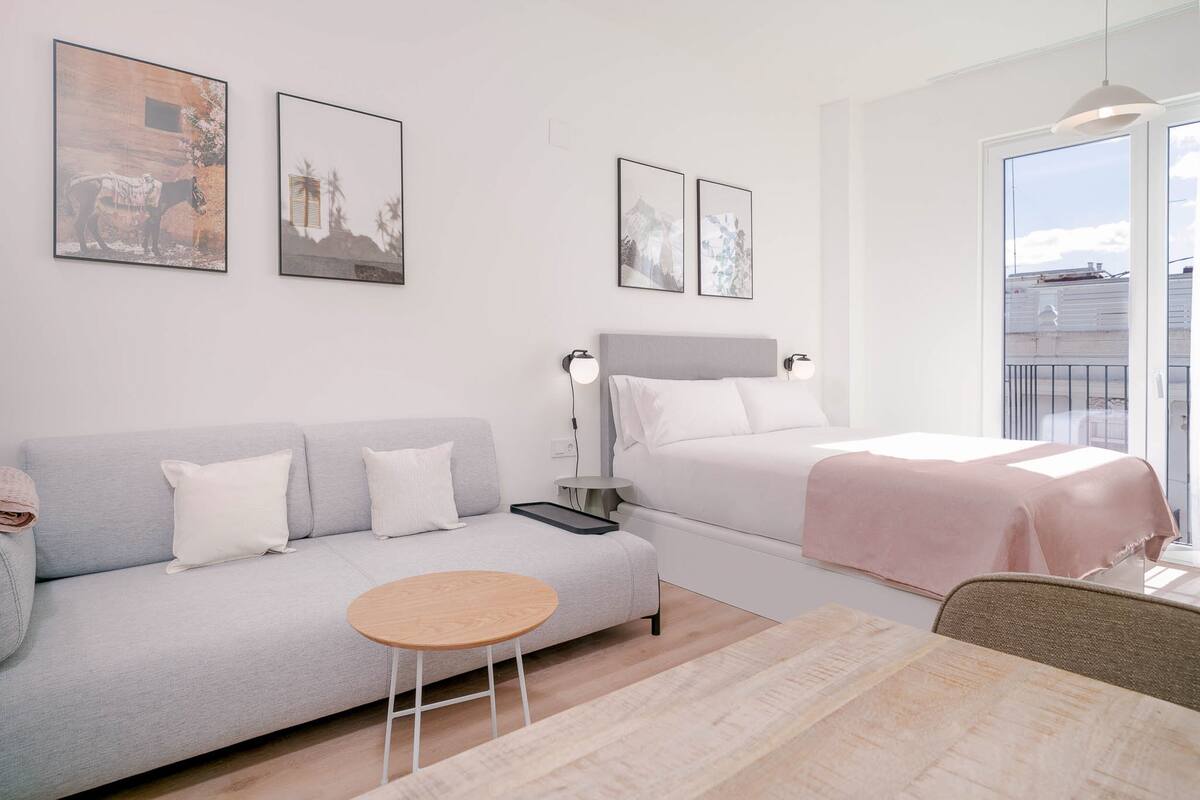 Un dormitorio moderno con un sofá gris, una cama individual con cabecera gris, una mesita redonda pequeña y dos obras de arte enmarcadas en la pared.