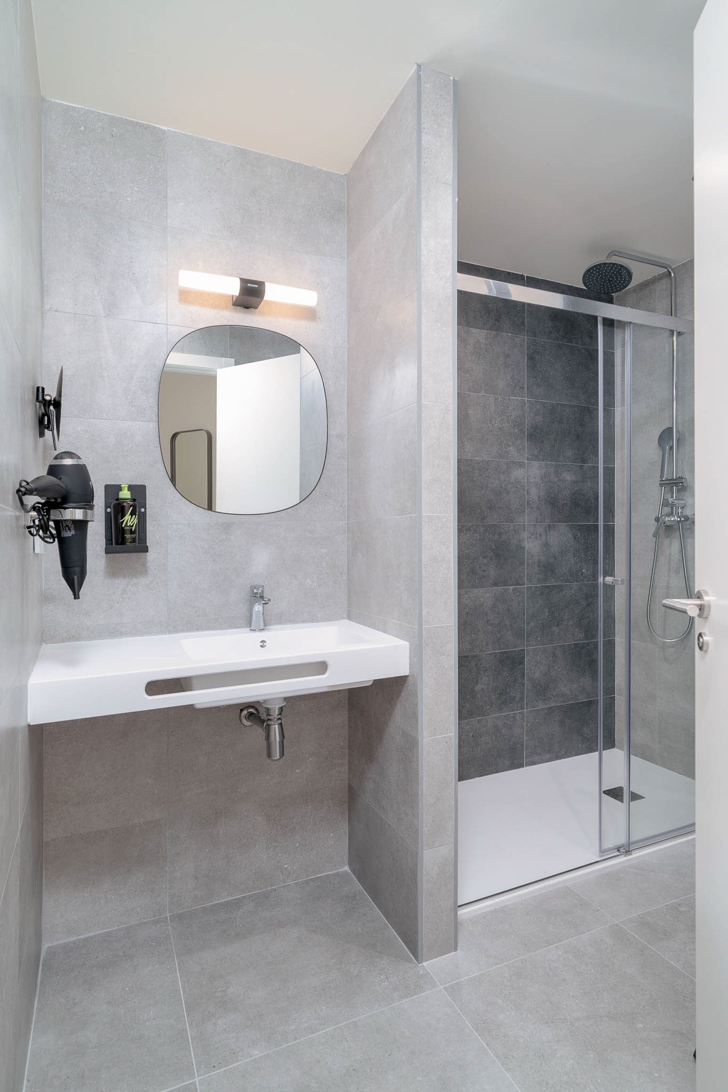 Un baño moderno con un lavabo blanco, un espejo redondo y una ducha con cerramiento de vidrio.