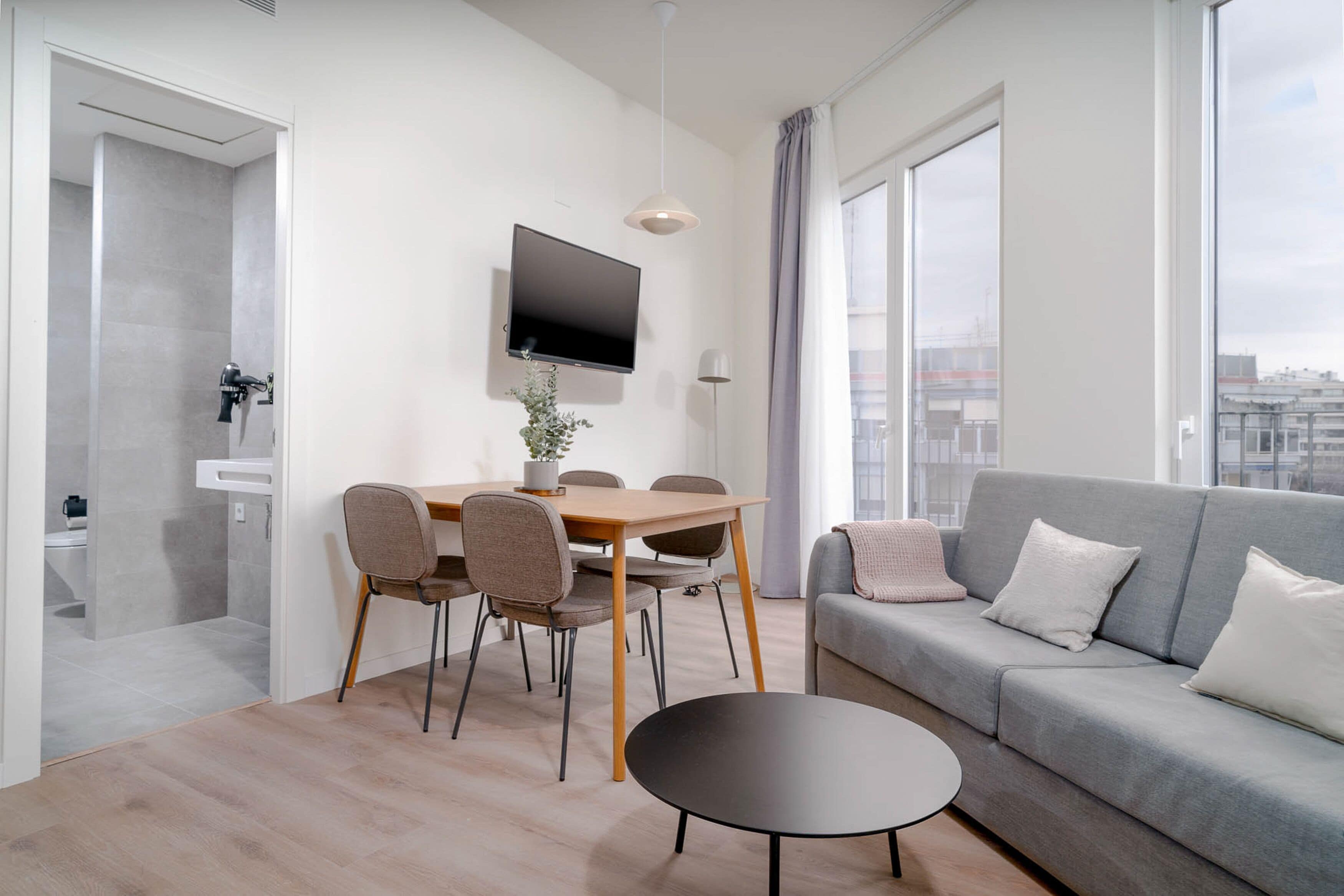 Un salon moderne avec un canapé gris, une table basse ronde et un coin repas comprenant une table et des chaises. Un téléviseur à écran plat est fixé au mur, et une grande fenêtre offre une vue sur la ville.