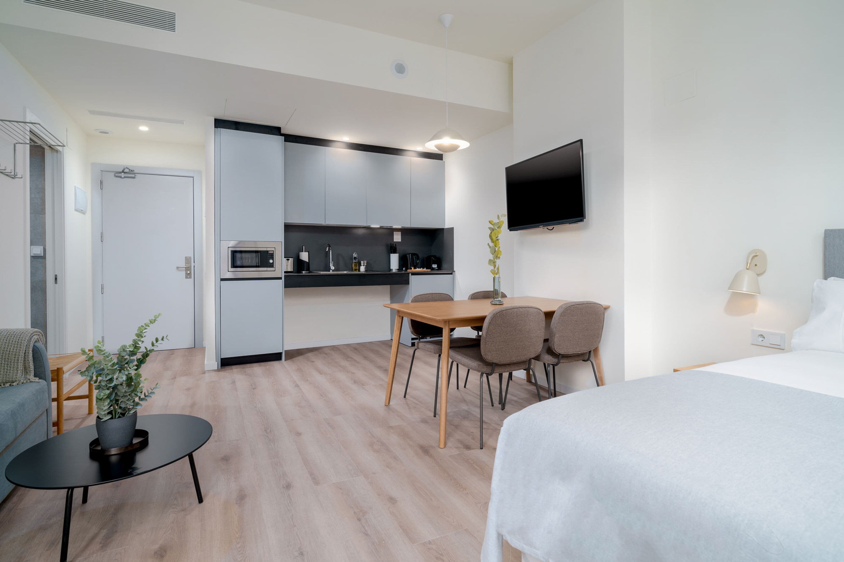 Une chambre d’hôtel moderne avec un lit, une table à manger avec des chaises, une petite table basse, un téléviseur fixé au mur et, au fond, une kitchenette.